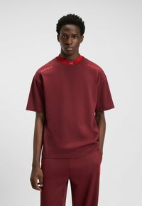 Bordeaux atletisk top med høj krave og røde detaljer, der har "HUGO" branding på skuldrene og en glat, blød tekstur.