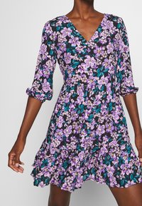 Blumenkleid vor einem dunklen Hintergrund mit lila und türkisfarbenen Blumen. V-Ausschnitt, Dreiviertelärmel und ein geraffter Saum. Baumwollmaterial.