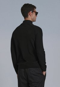 Schwarzer Pullover mit Hohem Kragen, Slim-Fit und gerippten Bündchen. Kombiniert mit dunklen Hosen, die eine glatte Stricktextur zeigen.