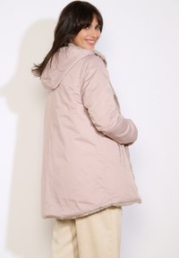 Giacca imbottita rosa chiaro con cappuccio, foderata in pelliccia, tasche laterali e chiusura con zip. Texture liscia con una finitura leggermente lucida.