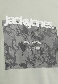 Světle zelené tričko s tmavě šedým abstraktním grafikou a bílým nápisem "jack&jones" a "CORE NINE ZERO." Hladká bavlněná textura.