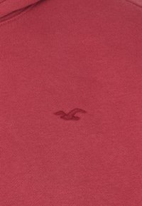 Sudadera roja con una textura suave y un pequeño logo bordado en la esquina inferior. Diseño simple y clásico sin adornos.