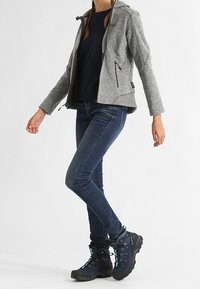 Osoba w szarej bluzie z zamkiem, ciemnoniebieskich dżinsach i niebieskich butach trekkingowych stoi z skrzyżowanymi nogami na białym tle.