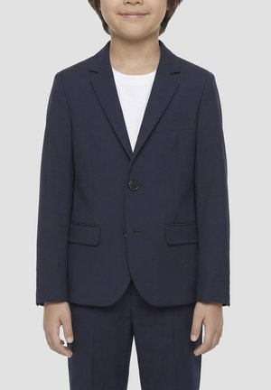 Bambino che indossa una giacca da abito blu scuro sopra una camicia bianca, in piedi con le braccia rilassate contro uno sfondo grigio uniforme.