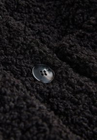 Black textured fabric with a fuzzy surface, featuring a round black button with two holes, positioned against a soft, thick background.