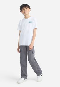 Boy standing wearing white Abercrombie & Fitch t-shirt, gray cargo pants, and white Nike sneakers with black swoosh on white background.