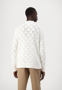 Maglione bianco lavorato a maglia con motivo a diamante, polsini e orlo a costine. Il modello indossa pantaloni beige. La vista posteriore mostra i dettagli a trama aperta.
