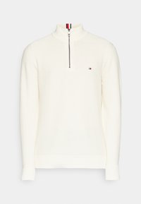 Bílý texturovaný pletený svetr s čtvrtinovým zipem, žebrovaným límcem a manžetami a malým logem v červené, bílé a modré barvě na hrudi.