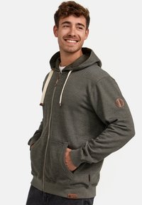 Sudadera gris con cremallera, fabricada en un tejido texturizado, con capucha con cordones, bolsillos frontales y detalle del logo en la manga. Estilo casual.