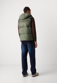 Green padded vest with a hood, worn over a brown long-sleeve shirt. Blue denim jeans and light sneakers complete the look from the back view.