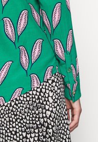 Camicia verde con motivo di foglie rosa e contorni blu, abbinata a una gonna nera con motivi a cuore bianchi. La camicia ha polsini con bottoni.