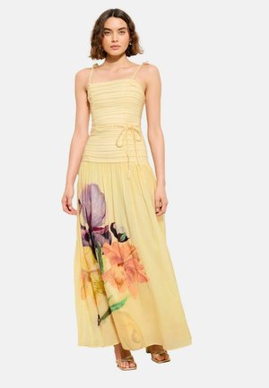 Kvinde iført en ærmeløs, lysegul maxi-kjole med blomsterprint på nederdelen og tynde skulderstropper bundet i sløjfer.