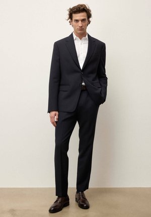 Homme portant un costume bleu marine ajusté avec une chemise blanche et des chaussures en cuir marron, debout avec une main dans la poche.