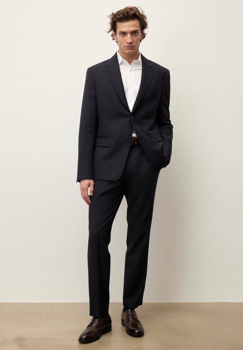 Homme portant un costume bleu marine ajusté avec une chemise blanche et des chaussures en cuir marron, debout avec une main dans la poche.