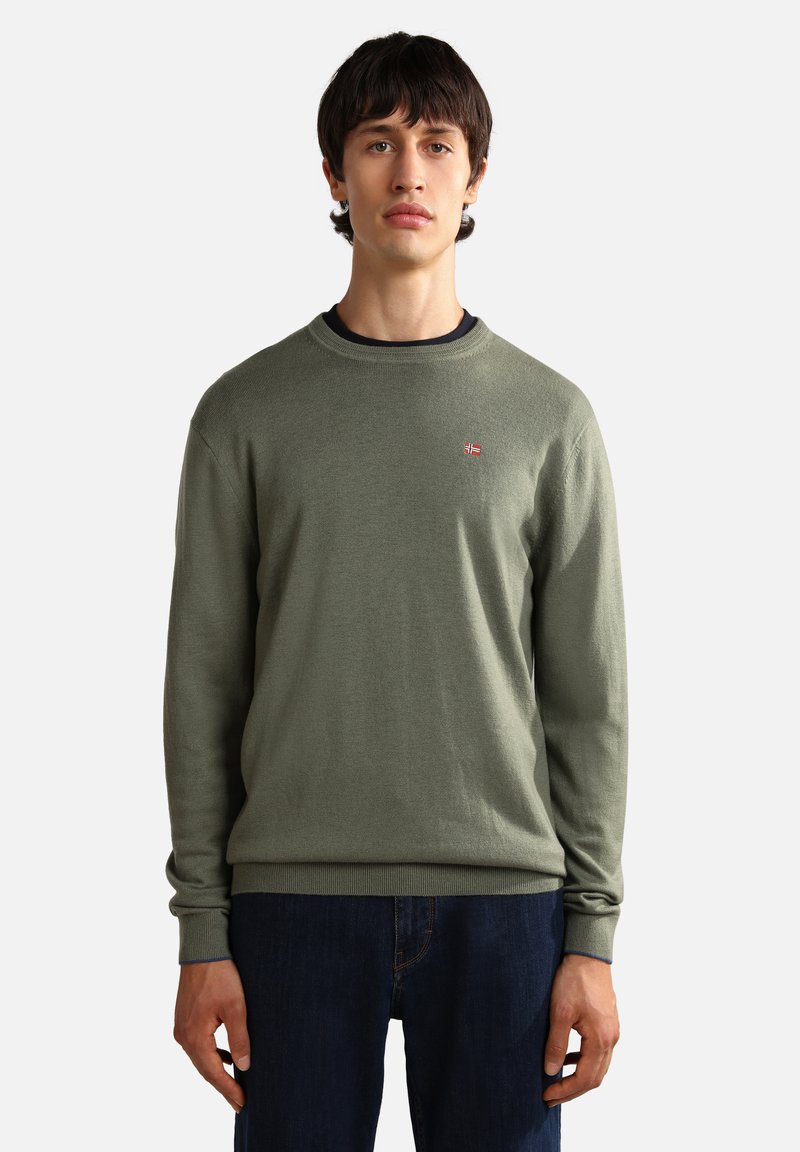Grüner Strickpullover mit Rundhalsausschnitt, versehen mit einem kleinen Flaggenemblem auf der Brust, kombiniert mit dunkelblauen Jeans. Weiche Textur, lässige Passform.