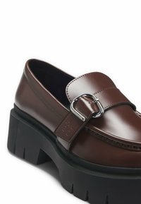 Mocassins en cuir marron à finition brillante, dotés d'une boucle argentée et d'une semelle plateforme noire épaisse avec un motif texturé.