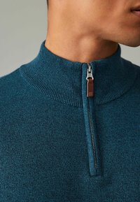 Türaqua Halb-Reißverschluss-Pullover mit geripptem Kragen, ausgestattet mit einem silbernen Reißverschluss und einem braunen Leder-Zugband. Strukturierter Stoff mit dezentem Muster.