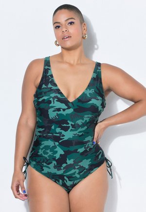 Žena s krátkými vlasy, která nosí zelené a černé maskáčové jednodílné plavky, zlaté kruhové náušnice a modrý lak na nehty, pózuje s rukou na bok.