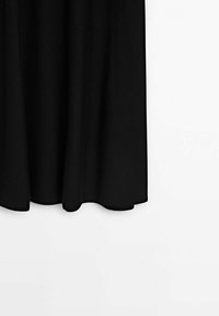 Ourlet de jupe en tissu noir suspendu sur fond blanc uni, montrant des plis doux et une texture lisse.