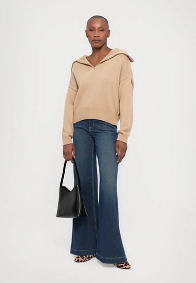 Hellbeige gestrickter Pullover mit Kapuze, kombiniert mit dunkelblauen Schlagjeans. Die Accessoires umfassen eine schwarze Tasche und Schuhe mit Animal-Print.
