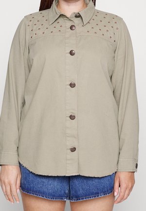 Chemise beige à manches longues boutonnée avec détail clouté aux épaules portée sur un short en denim bleu, sur une personne debout devant un fond uni.