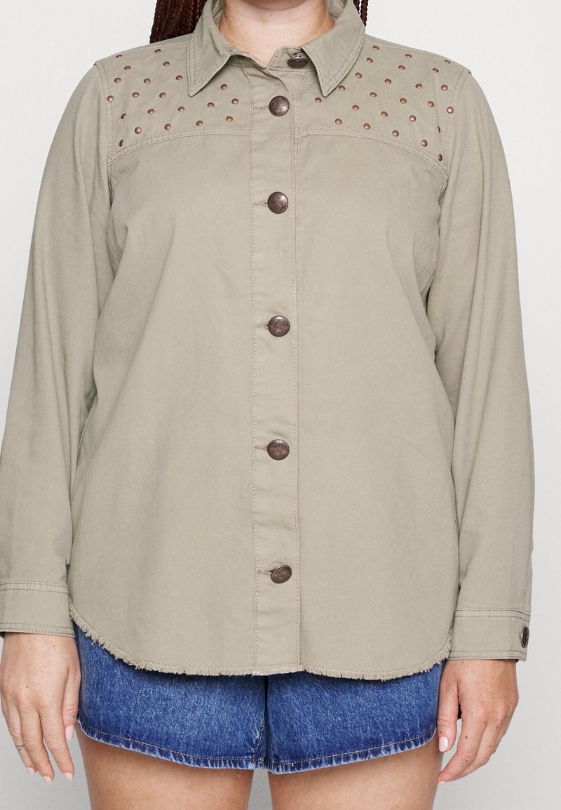 Chemise beige à manches longues boutonnée avec détail clouté aux épaules portée sur un short en denim bleu, sur une personne debout devant un fond uni.