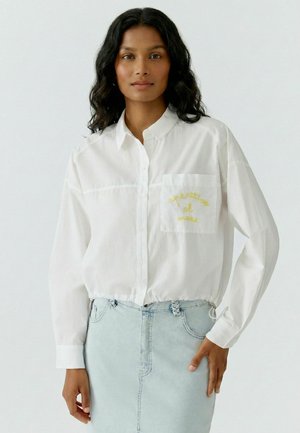 Femme aux cheveux ondulés foncés portant une chemise blanche boutonnée avec un texte jaune sur la poche et une jupe en jean bleu clair.