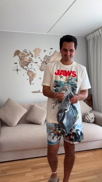 Μαύρο αντικείμενο σε διαφανή σακούλα που κρατά ένα άτομο ντυμένο με μπλούζα γραφικών JAWS και σορτς με παλμό παλμικών φύλλων, standing σε ένα ανοιχτόχρωμο δωμάτιο.