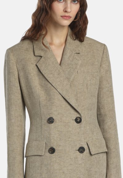 Femme portant un blazer beige croisé à larges revers avec quatre boutons noirs, assorti de poches à rabat.