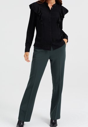 Kobieta ubrana w czarną bluzkę z długim rękawem i marszczonymi ramionami oraz ciemnozielone spodnie o prostych nogawkach, stojąca z jedną ręką w kieszeni.