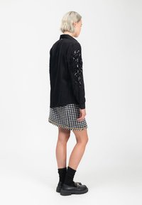 Schwarzes Langarmshirt mit strukturierten Verzierungen an den Ärmeln, kombiniert mit einem Hahnentrittrock mit goldakzentuiertem Saum. Schwarze Schuhe.