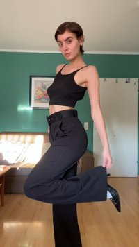 Top cropped preto e calças pretas de cintura alta com um cinto. O modelo posa com uma perna levantada, destacando o design elegante da calça.