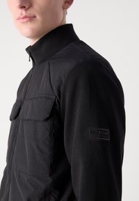 Jachetă neagră cu guler înalt, material texturat, închidere cu fermoar și două buzunare pe piept; are o etichetă logo neagră pe mânecă.