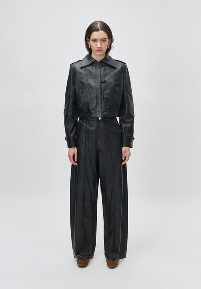 Νεαρός ενήλικας που φοράει μαύρο κοντό δερμάτινο μπουφάν και δερμάτινο παντελόνι φαρδιάς γραμμής, στέκεται μπροστά σε απλό, φωτεινό φόντο.