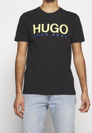 Camiseta de algodón negra con texto en negrita amarillo y azul: "HUGO" arriba y "HUGO BOSS" debajo. Cuello redondo clásico, mangas cortas.