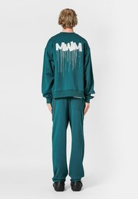 Sudadera oversized de color verde azulado con texto blanco goteante en la parte posterior, combinada con pantalones ajustados a juego y zapatos negros de suela gruesa.