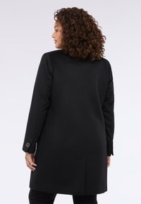 Abrigo negro largo con una silueta recta, cuello con muesca y detalles de botones en las mangas. Tejido suave, con una abertura sencilla en la parte posterior.
