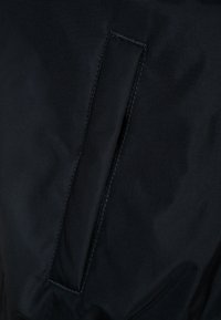 Černá textilní kapsa s obdélníkovým otvorem, s hladkým povrchem a čistým švem, integrovaná do většího oděvu.