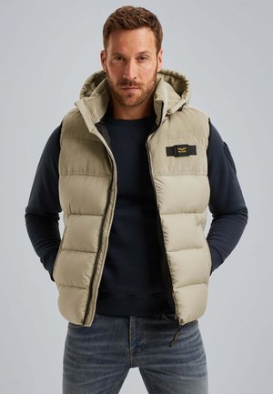 Mann trägt eine beige gepolsterte ärmellose Weste über einem schwarzen Langarmshirt und blauen Jeans, steht mit den Händen in den Taschen der Weste.