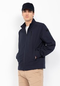 Veste zippée marine avec col montant, poches latérales et poche sur la poitrine ; fabriquée en tissu léger ; associée à une casquette assortie.