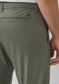 Olivgrüne Baumwollmischungshose mit einer flachen Gesäßtasche und einem kleinen Logodetail. Glatte Textur, taillierte Passform und sichtbare Seitentasche.