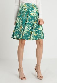 Una persona está de pie vistiendo una falda floral en tonos verdes y sandalias de tacón nude, con un suéter blanco de canalé, frente a un fondo liso.