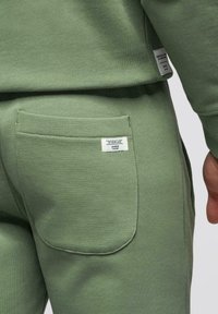 Gros plan sur une personne portant un pantalon de survêtement vert et un sweat-shirt, montrant une poche arrière avec une étiquette rectangulaire blanche sur le pantalon.