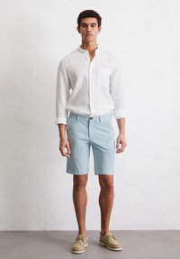 Chemise boutonnée en lin blanc, shorts bleu clair et chaussures beiges. Le modèle se tient devant un fond neutre, mettant en valeur une tenue d'été décontractée.
