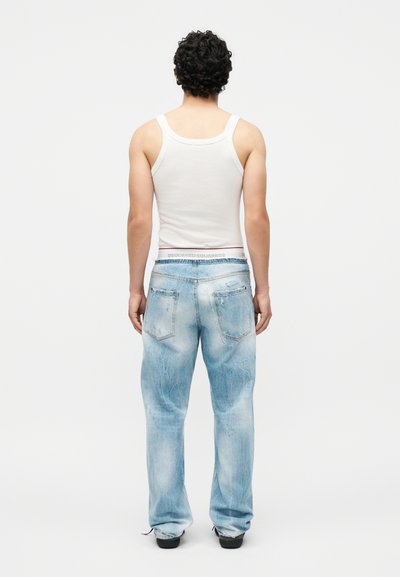 Άνδρας που στέκεται γυρισμένος με την πλάτη, φοράει λευκό φανελάκι και ανοιχτό μπλε ξεβαμμένο τζιν με μαύρα παπούτσια, σε απλό λευκό φόντο.