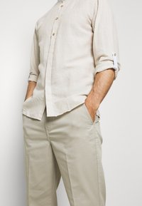 Camisa de lino beige clara con botones en la parte delantera y mangas enrolladas, combinada con pantalones beige claros de textura suave y corte recto.