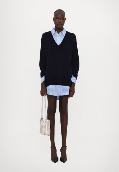 Marineblauer Oversize-Pullover mit V-Ausschnitt, über einem hellblau gestreiften Hemd getragen. Das Model hält eine kleine, gewebte beigefarbene Handtasche und trägt schwarze High Heels.