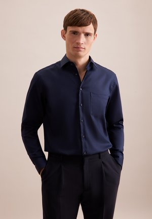 Chemise à manches longues marine avec col boutonné, poche poitrine et boutons sur le devant. Tissu lisse avec une coupe ajustée. Pantalons foncés.