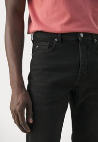 Homme portant un jean noir et un t-shirt rose saumon, debout avec son bras gauche relâché le long du corps devant un fond uni.