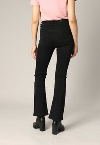 Jean flare noir à taille haute, avec poches arrière et détails cousus. Associé à un haut rose clair à manches courtes et des talons épais noirs.